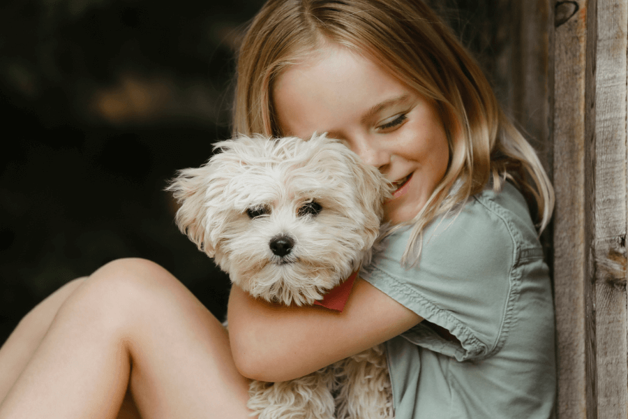 Smiling young girl with long blonde hair cuddling a fluffy white dog in a relaxed, outdoor setting, expressing a moment of affection and companionship.