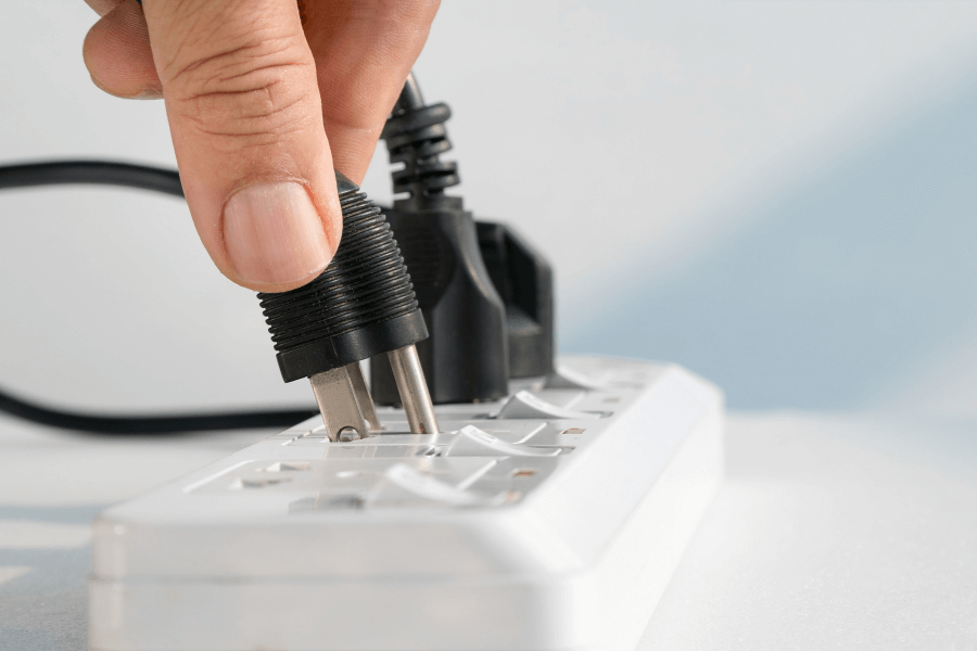 This image shows a hand plugging a black electrical plug into a white power strip. The setup suggests a practical, everyday action, with the power strip already containing a few connected plugs. The clean, minimal background emphasizes the focus on the hand and the plug.