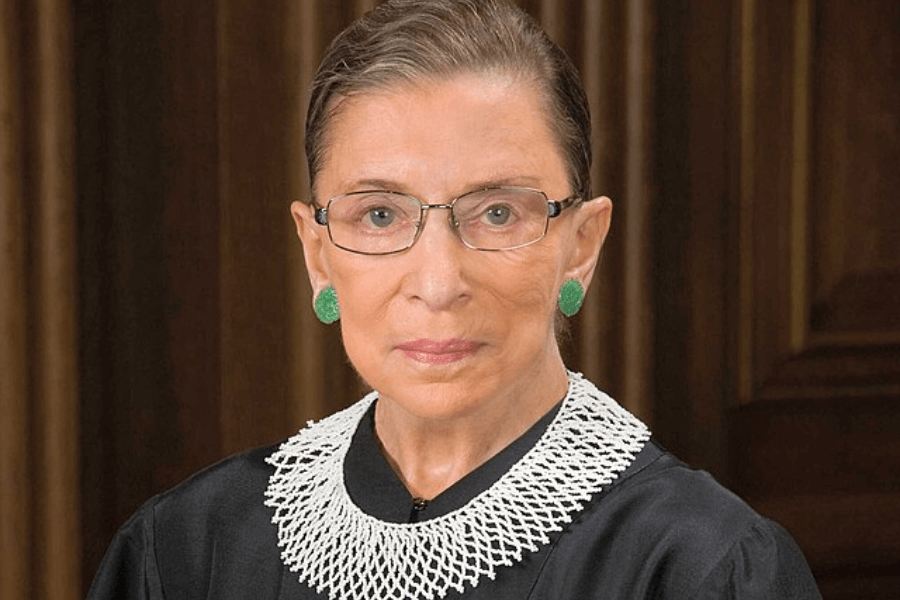 A formal portrait of a woman wearing a black judicial robe with a distinctive white lace collar. She has neatly pulled-back hair, glasses, and green earrings, with a composed and serious expression. The background features dark wooden paneling, enhancing the dignified setting.