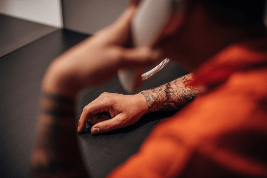 Tattooed man in an orange jumpsuit speaking on a prison visitation phone, with only his arm and part of his face visible, highlighting a moment of communication from incarceration.