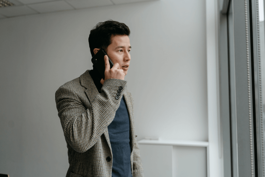 A man in a checkered blazer and dark t-shirt is standing near a large window, engaged in a phone call. His expression appears focused, and the modern, minimalist office setting enhances the professional atmosphere.