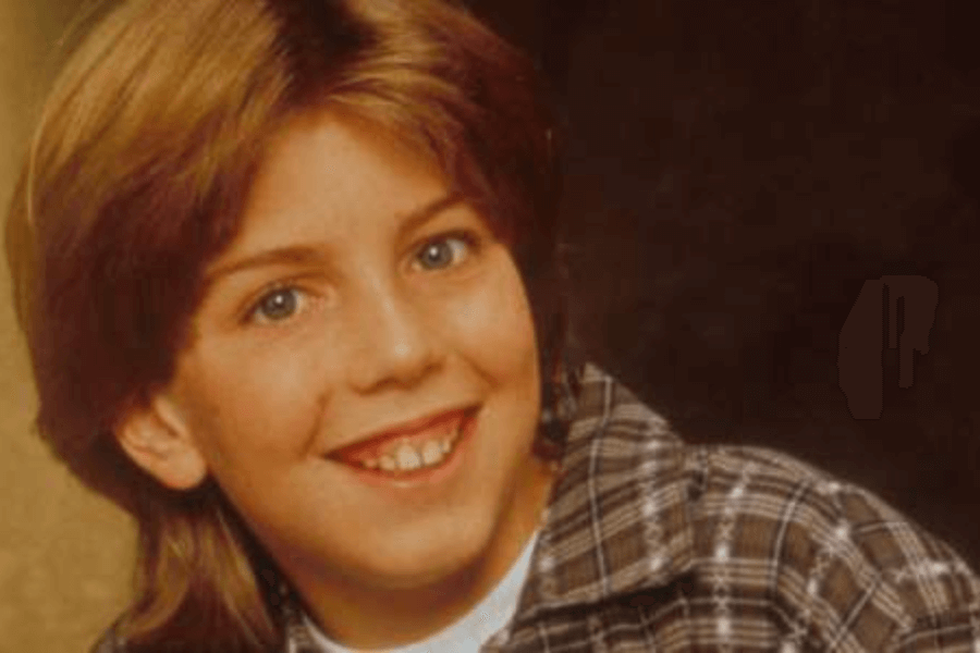 This image features a young boy with light brown hair styled in a classic 90s haircut, smiling brightly. He is wearing a plaid shirt, and the background is softly lit, giving a warm, nostalgic portrait feel.
