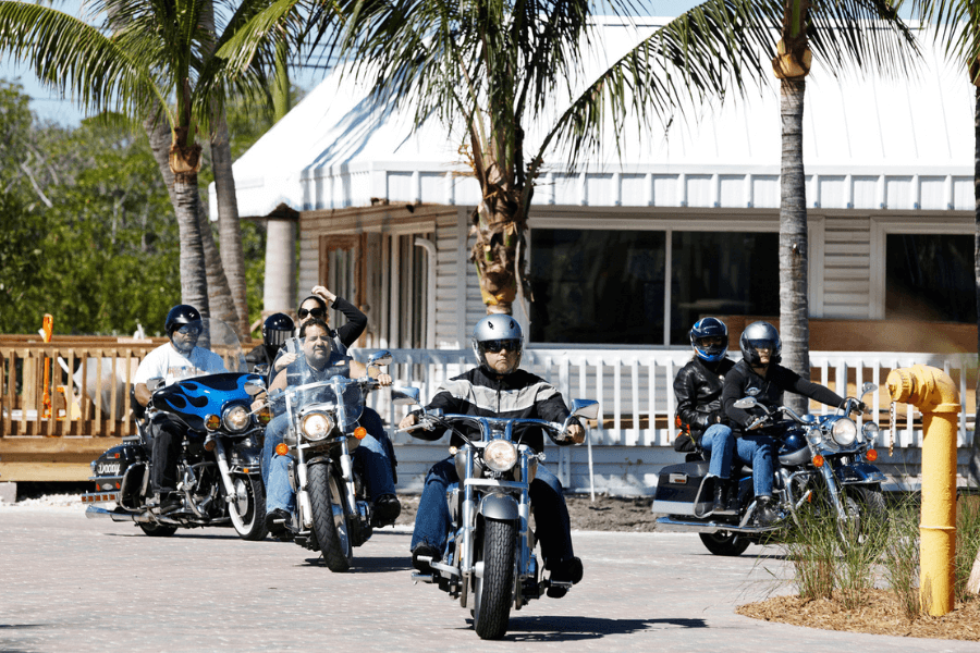 Group of motorcyclists riding cruisers on a sunny day near palm trees and a white building, with some wearing helmets and leather jackets, capturing a sense of freedom and camaraderie.