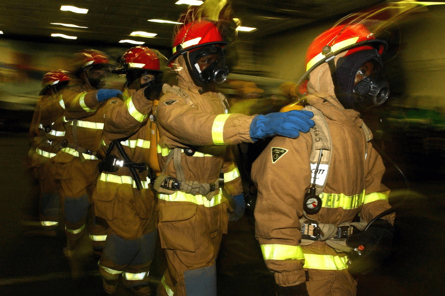 This image shows a team of firefighters in full protective gear, including red helmets and gas masks, moving in a coordinated line. Each firefighter rests a hand on the shoulder of the person in front, emphasizing teamwork and visibility in a potentially hazardous environment. The blurred background and lighting suggest a sense of urgency or action.