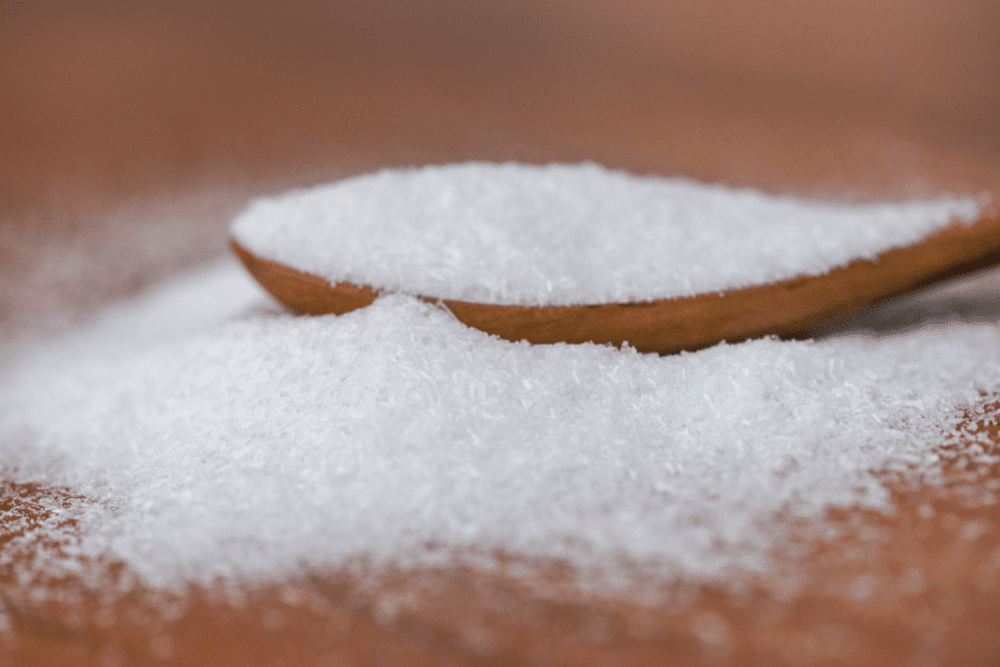 A close-up of a wooden spoon filled with white crystalline powder, likely monosodium glutamate (MSG), resting on a wooden surface. The fine granules spill over, forming a small pile around the spoon.
