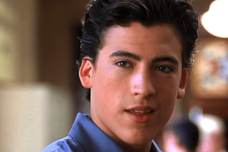 This image shows a young man with neatly styled dark hair and a confident expression. He is wearing a light blue shirt, and the background features a softly blurred indoor setting, giving the image a polished and warm tone.
