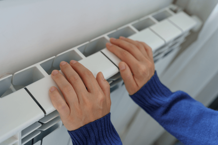 This image shows two hands resting on a white radiator, suggesting someone is warming their hands. The individual is wearing a blue sweater, and the setting appears to be indoors with a focus on the radiator's structure.