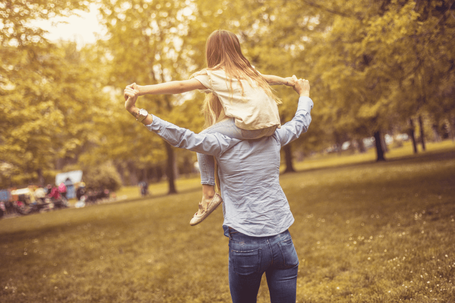 Father carrying his young daughter on his shoulders in a sunlit park, both holding hands and enjoying a playful moment surrounded by trees and soft golden light.