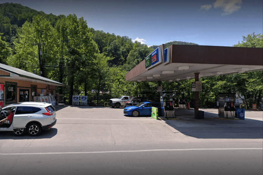 A small gas station with a brown canopy and multiple fuel pumps is situated in a rural, tree-lined area. A convenience store named "C & L" is adjacent to the gas station, with signs and coolers displayed outside. Several vehicles, including a blue car at the pumps and a white SUV with an open door, are present in the lot.