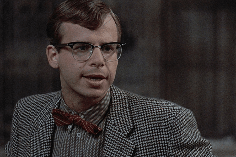 This image features a man wearing glasses, a checkered blazer, a striped shirt, and a red bow tie. He has a thoughtful expression and is captured in what appears to be an indoor setting, with a neutral or dimly lit background.