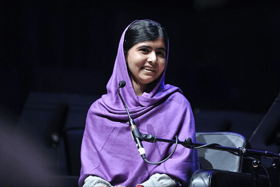 A young woman wearing a purple headscarf sits on a stage, smiling while speaking into a microphone. She appears confident and engaged, with a dark background and stage lighting illuminating her face. A microphone stand is positioned in front of her, suggesting she is participating in a discussion or giving a speech.
