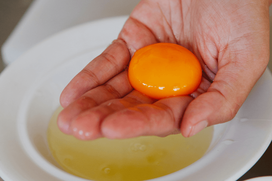 This image shows a close-up of a hand holding a vibrant orange egg yolk, carefully separated from the egg white visible in a bowl underneath. The focus highlights the delicate and glossy texture of the yolk.