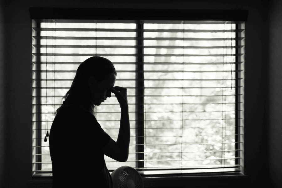 Black and white silhouette of a person standing in front of a window with blinds, holding their forehead with a tense or thoughtful expression, evoking a sense of stress, fatigue, or contemplation.