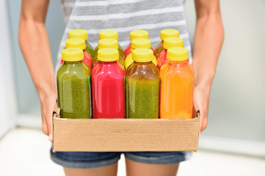 A person in a striped shirt and denim shorts holds a cardboard box filled with colorful bottled juices. The bottles, sealed with yellow caps, contain vibrant green, red, and orange smoothies or juices.