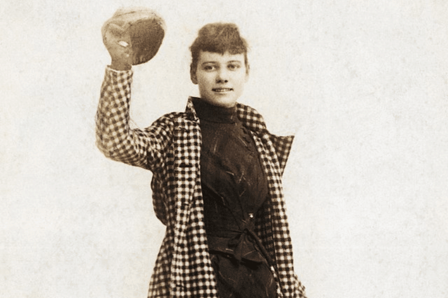 A vintage sepia-toned photograph of a woman wearing a checkered coat over a dark dress, holding up a round hat with a confident smile. Her short, wavy hair and relaxed pose give her an adventurous and independent demeanor. The plain background emphasizes her as the focal point of the image.