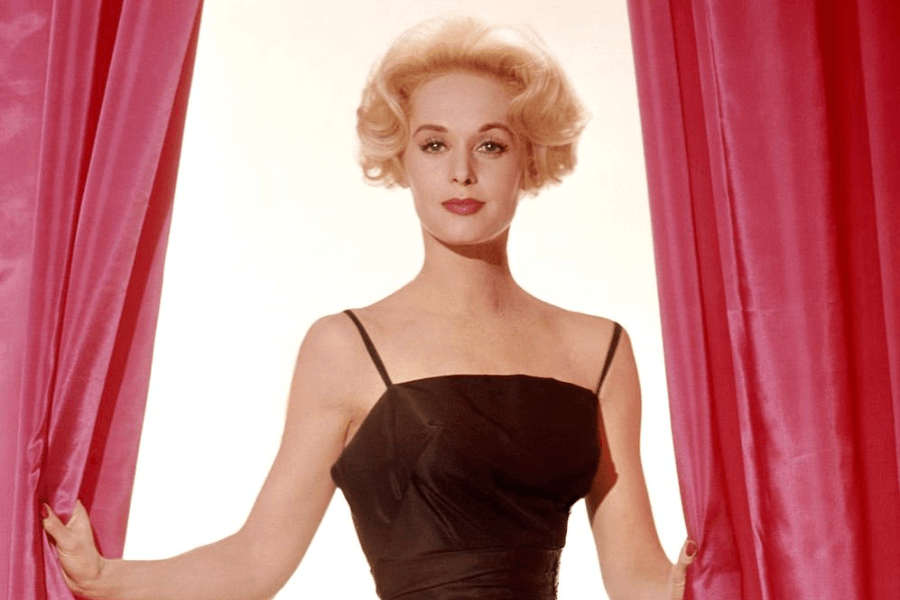 This image features a woman with short, styled blonde hair, wearing a classic black dress with thin straps. She is standing between two vibrant pink curtains, holding them open, with a poised and elegant expression. The bright, minimalist background emphasizes her timeless and sophisticated look.
