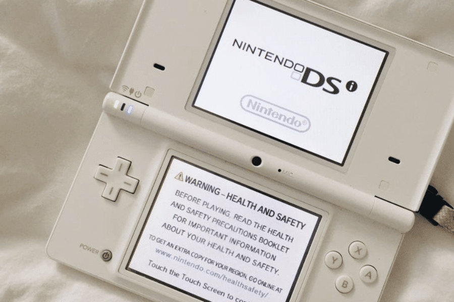This image shows a white Nintendo DSi handheld gaming console, opened and powered on. The top screen displays the "Nintendo DSi" logo, while the bottom screen shows a health and safety warning. The console rests on a light fabric surface, giving it a casual and relaxed setting.