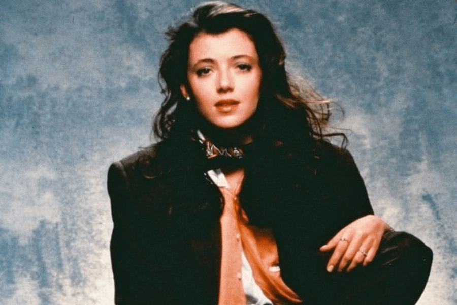 This image features a woman with long, wavy dark hair, wearing a formal yet eclectic outfit that includes a suit jacket, a peach-colored blouse, and a decorative scarf around her neck. She has a relaxed and confident pose, with one hand resting on her knee, set against a textured blue-gray backdrop that emphasizes a vintage and sophisticated aesthetic.