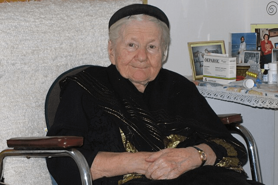 An elderly woman with short white hair sits in a wheelchair, wearing a black outfit with gold embroidery and a black headpiece. She has a gentle expression with folded hands resting in her lap. Behind her, a table with medications, framed photographs, and personal items suggests a cozy and familiar setting.