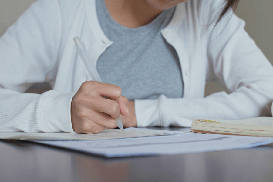 This image shows a person sitting at a desk, writing with a pen on a sheet of paper. They are wearing a white cardigan over a gray shirt, and an open notebook is visible nearby. The scene conveys focus and engagement, likely related to studying or note-taking.