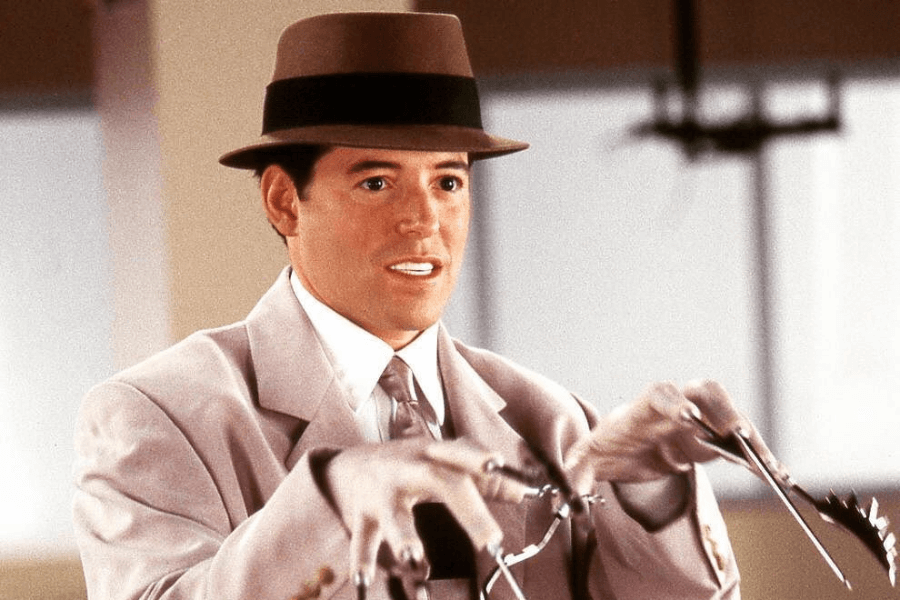 This image features a man wearing a beige suit, a tie, and a brown fedora, evoking a classic or retro style. He appears to be operating or examining a mechanical device, with a slightly surprised or focused expression. The setting is indoors with soft lighting, emphasizing a polished and vintage aesthetic.