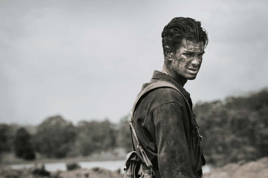 Scene from a war film showing a soldier covered in dirt and grime, wearing a worn military uniform and gear, standing against a desolate, cloudy backdrop. His intense expression and battle-worn appearance reflect the emotional toll of combat.