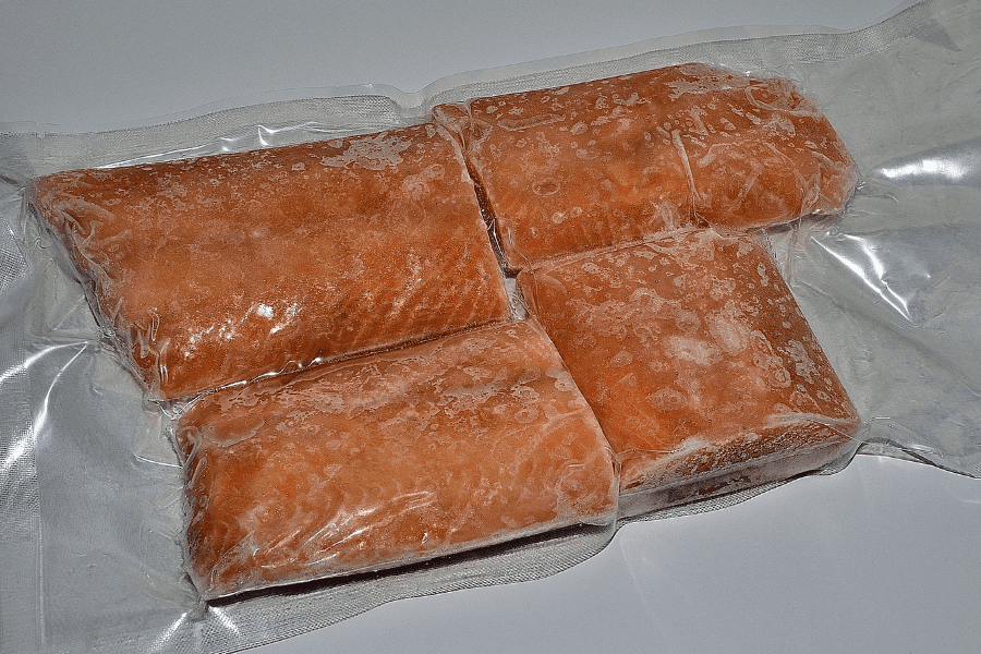 This image shows four frozen salmon fillets vacuum-sealed in clear plastic packaging. The frost on the surface indicates they are well-preserved and ready for storage or use. The simple white background emphasizes the focus on the food product.