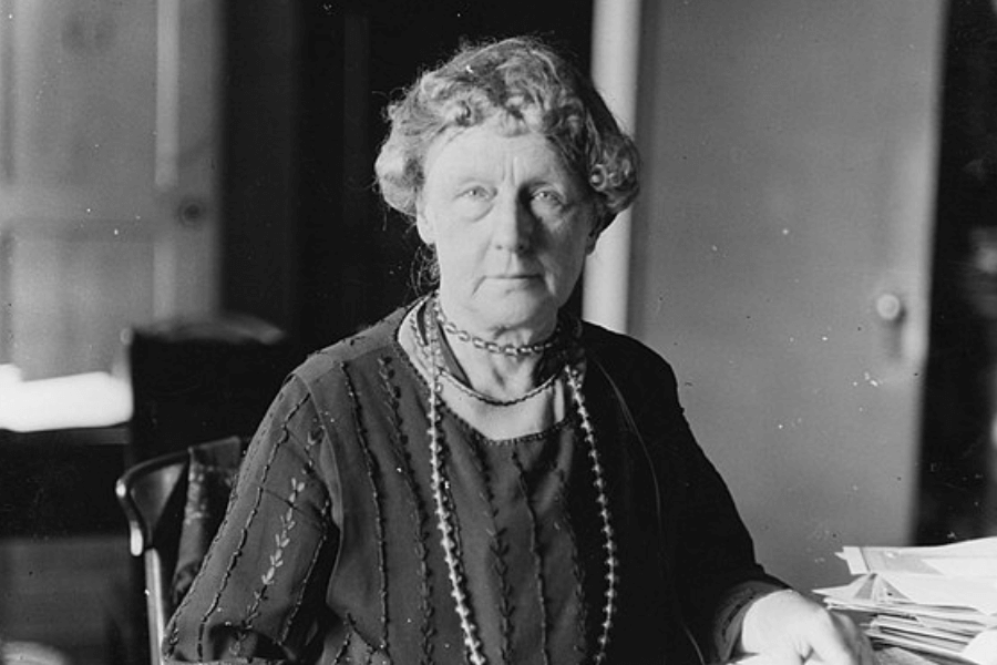 A black-and-white photograph of an older woman seated at a desk with a serious and focused expression. She wears a dark embroidered dress, a pearl necklace, and a beaded choker, with her curly hair neatly styled. A stack of papers rests on the table beside her, suggesting a scholarly or administrative setting.