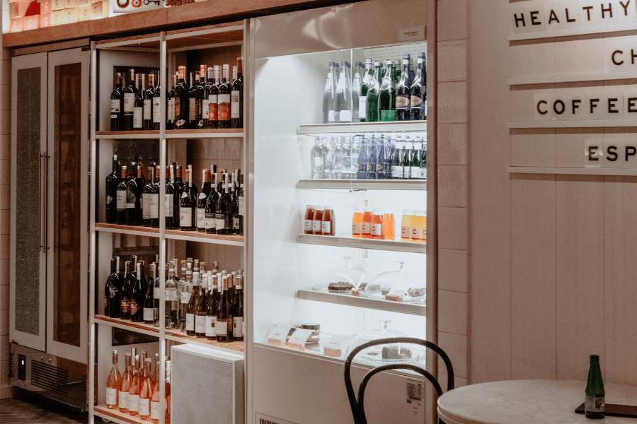 A display of wine bottles arranged on shelves next to a refrigerated case holding sparkling water, juices, and desserts. The setting is a modern café or wine bar, with minimalist decor and a letterboard menu on the wall.