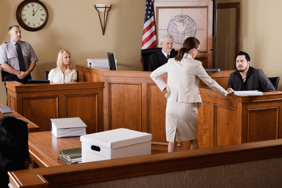 Female attorney questioning a male witness on the stand in a courtroom, with a judge, jury, and security officer present, alongside visible legal documents and evidence boxes.