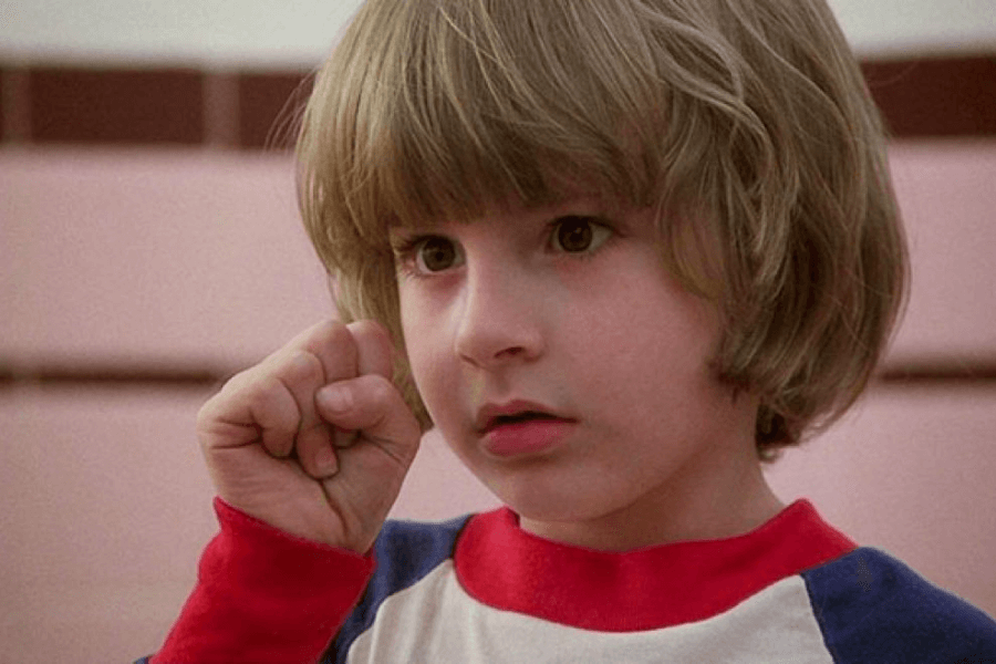 This image shows a young child with medium-length blonde hair and a focused expression, holding up a fist near their face. They are wearing a red, white, and blue shirt, standing in what appears to be an indoor setting with light pink and brown tiled walls. The image conveys a sense of curiosity or thoughtfulness.
