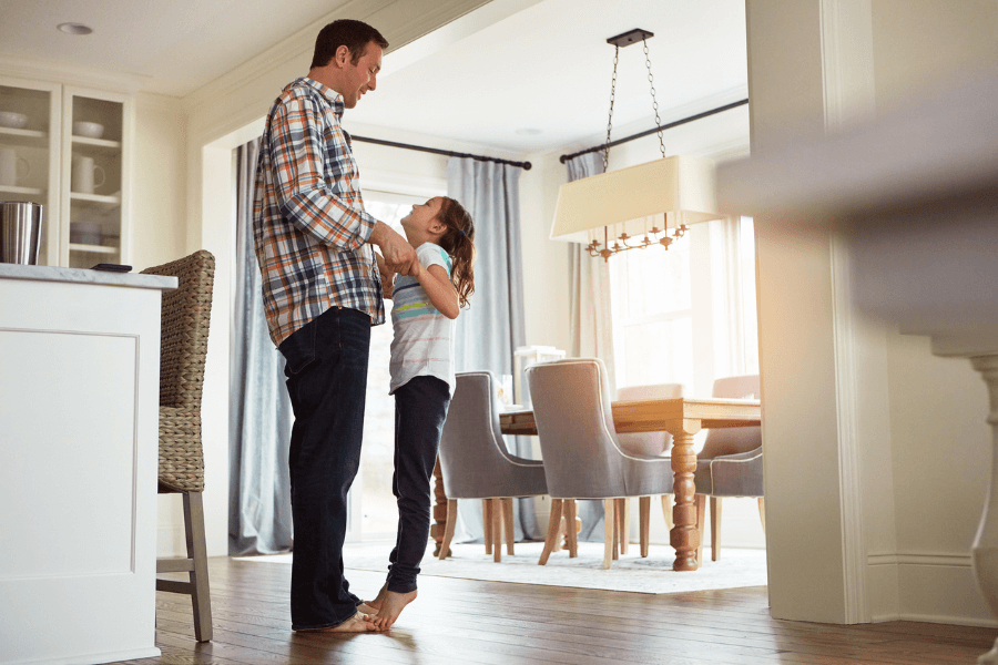 Father and young daughter sharing a happy moment in their kitchen, smiling at each other while holding hands, with sunlight streaming into a warmly decorated dining area.
