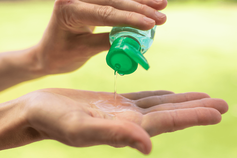 A close-up of a person pouring clear hand sanitizer from a small bottle with a green flip-top cap into their palm. The background is blurred and green, suggesting an outdoor setting.