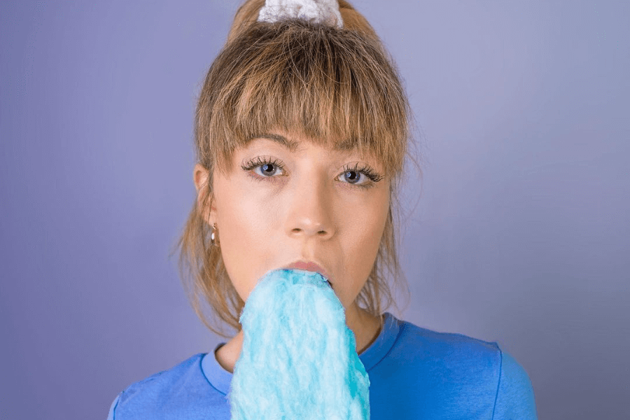 This image features a young woman with blonde hair styled in a ponytail with a scrunchie, wearing a blue shirt. She is holding and eating blue cotton candy against a light purple background. Her expression is neutral, and the bright colors give the image a playful and whimsical vibe.