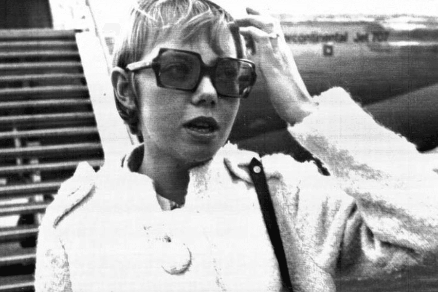 Black and white photo of a woman in oversized sunglasses and a textured coat, standing in front of an airplane staircase with part of the aircraft visible behind her. She is adjusting her hair and appears to be speaking or mid-sentence.