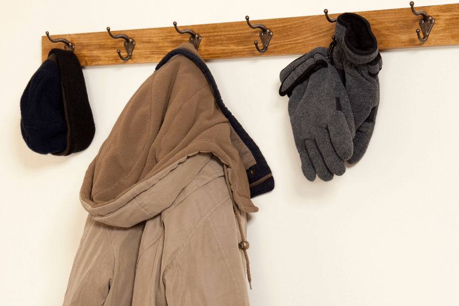 This image shows a wooden coat rack mounted on a wall, holding a beige jacket, a pair of gray gloves, and a dark winter hat. The hooks are evenly spaced, and the arrangement suggests a functional and organized entryway setup for cold-weather attire.