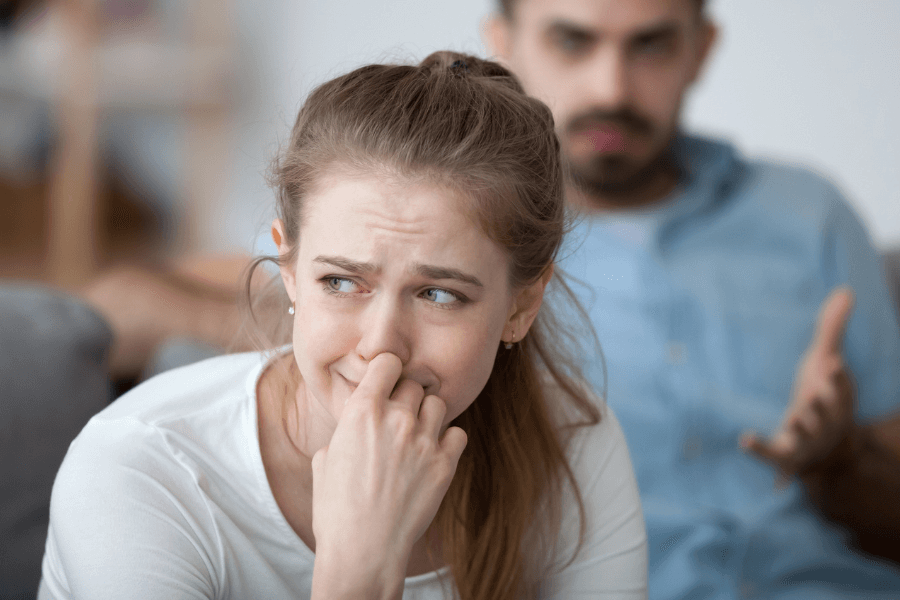 Young woman sitting on a couch with a distressed expression, holding her hand to her face, while a man in the background appears to be arguing or expressing frustration.