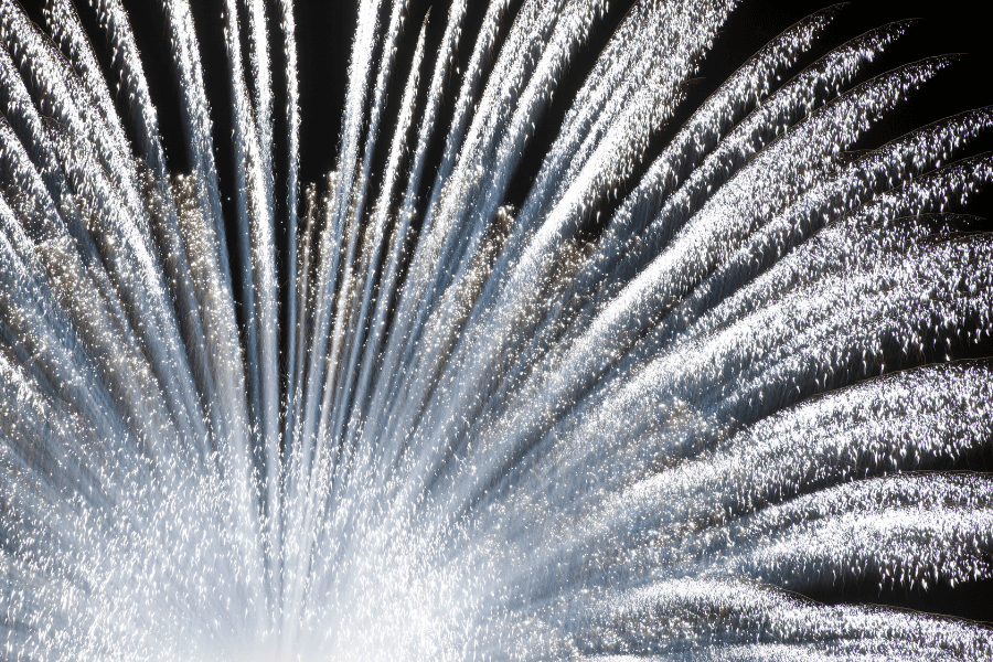 This image captures a stunning display of fireworks, with white and silver streaks radiating outward in an impressive fan shape against a dark night sky. The sparkling bursts create a dazzling and celebratory atmosphere.