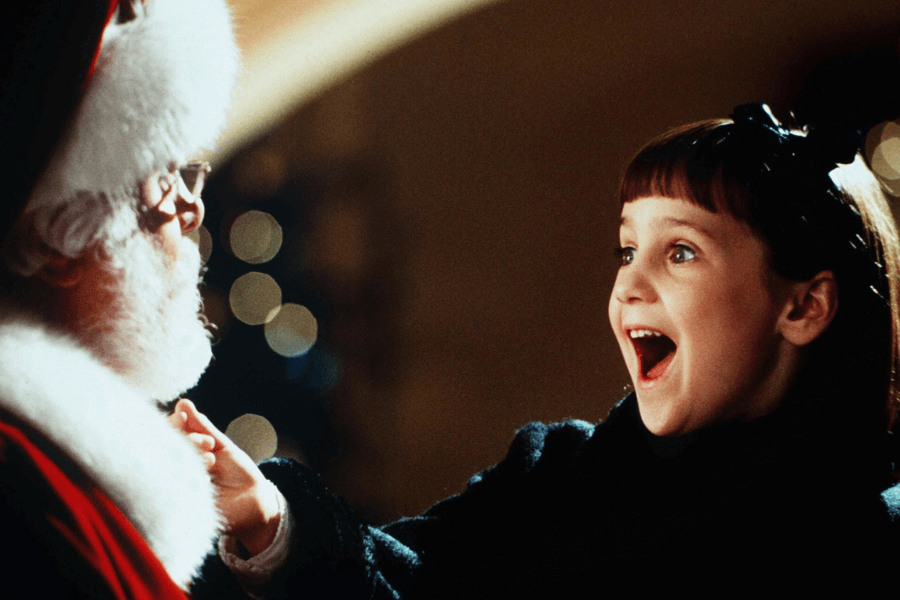 This image shows a young girl with an excited and joyful expression, reaching out toward a man dressed as Santa Claus. The scene is warmly lit with blurred Christmas lights in the background, creating a festive and magical holiday atmosphere.