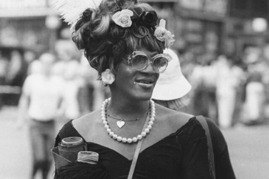 A black-and-white photograph of a person with a confident smile, wearing oversized sunglasses, a pearl necklace, and a heart-shaped pendant. Their voluminous, curled hair is adorned with flowers and a feather, and they are dressed in an off-the-shoulder top while holding a beverage can. The blurred background suggests a lively outdoor gathering or parade.