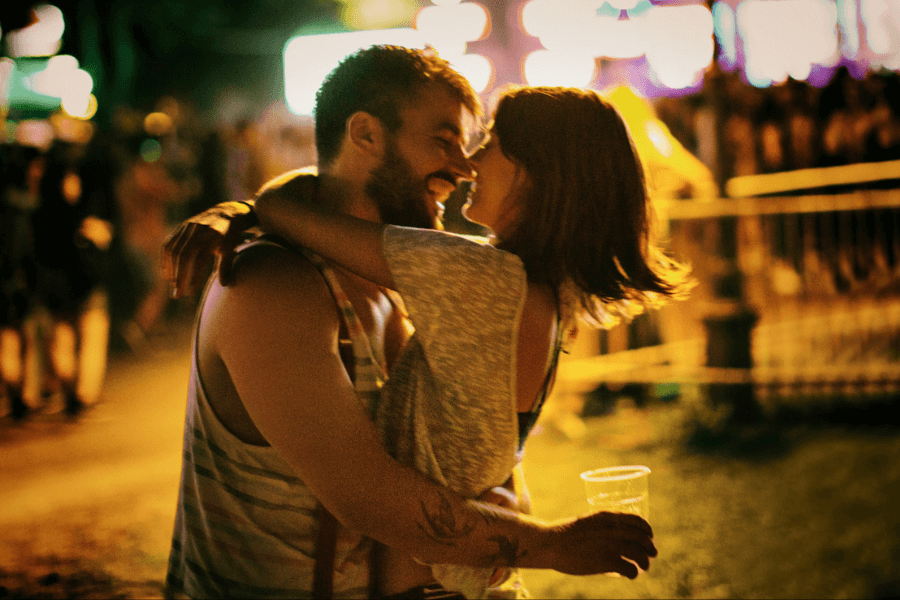 Young couple embracing and laughing under glowing festival lights at night, with a crowd in the background and a joyful, carefree atmosphere.