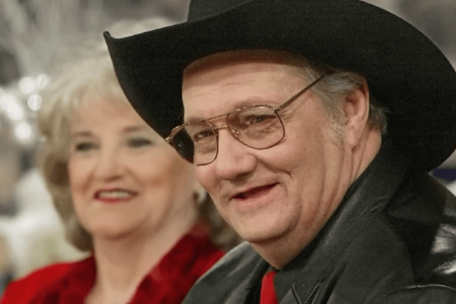 A man wearing a black cowboy hat, glasses, and a leather jacket smiles while looking slightly off-camera. Behind him, a woman dressed in red with short, wavy blonde hair is also smiling. The background is blurred, suggesting an indoor festive or formal event.
