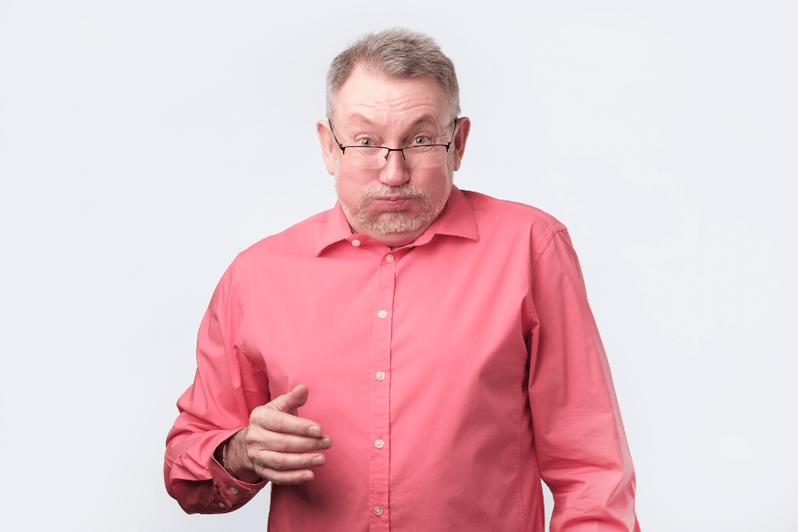 A middle-aged man wearing a pink button-up shirt and glasses makes a puffed-cheek expression, as if holding his breath or reacting to something unpleasant. His eyebrows are slightly raised, and he appears to be in a humorous or exaggerated moment.