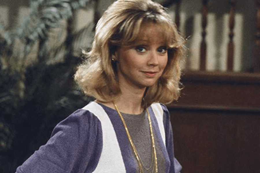 This image features a woman with shoulder-length blonde hair styled in soft waves, wearing a purple top layered with a white vest and accessorized with long gold necklaces. She is standing in an indoor setting with wooden railings and greenery in the background, giving a warm and classic vibe. Her expression is poised and friendly.