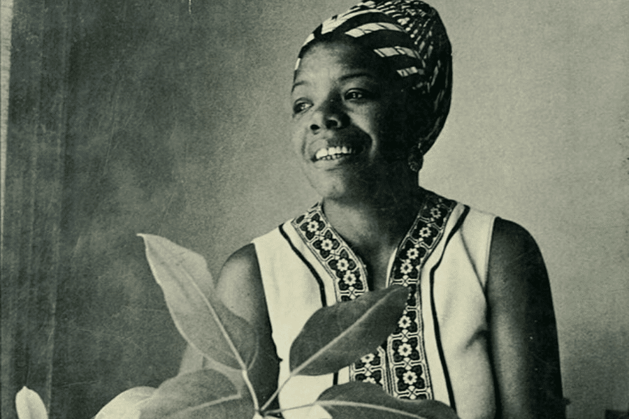 A black-and-white portrait of a woman wearing a patterned headwrap and a sleeveless embroidered top, smiling warmly. She is seated near a plant, with soft lighting casting gentle shadows on her face. The photograph captures a serene and expressive moment.