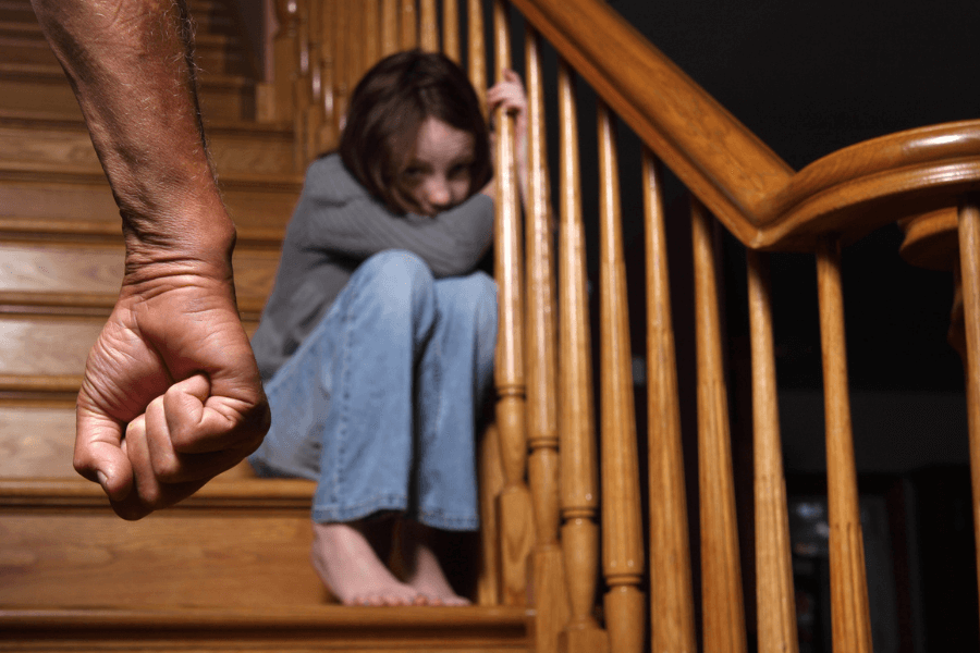 Frightened young girl sitting on a staircase, hugging her knees with a fearful expression, while an adult’s clenched fist is seen in the foreground, suggesting a threatening or abusive situation.