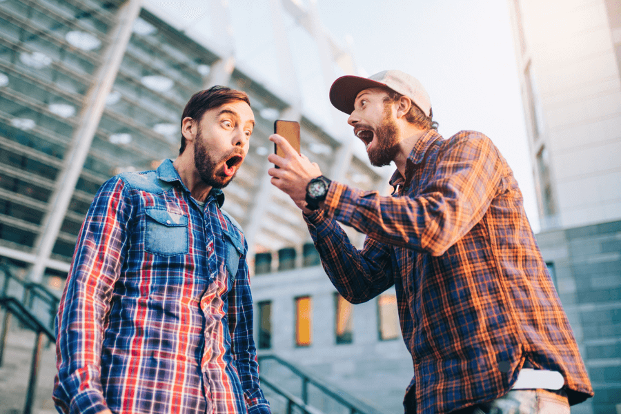 Two young men wearing plaid shirts and beards react with excitement and surprise while looking at a smartphone screen. One man, wearing a cap, holds the phone and appears to be sharing shocking news, while the other, dressed in a denim-accented plaid shirt, has his mouth wide open in astonishment. The background features modern architecture with glass and steel elements, suggesting an urban setting.