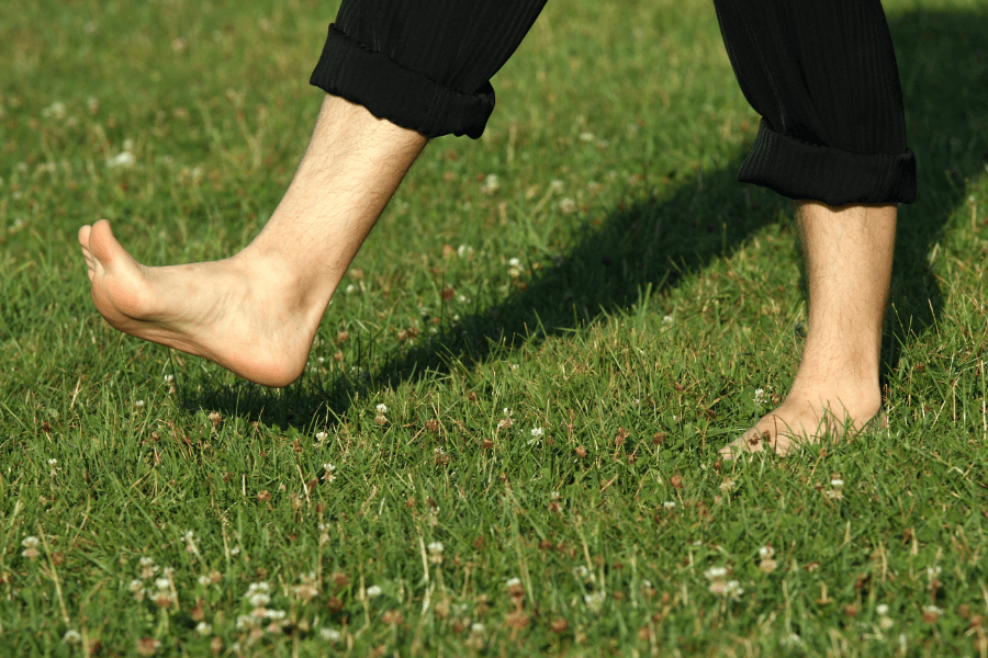 A close-up of a person's bare feet walking on a grassy field, with one foot lifted mid-step. The individual is wearing black pants rolled up slightly, and the green grass is dotted with small white flowers.