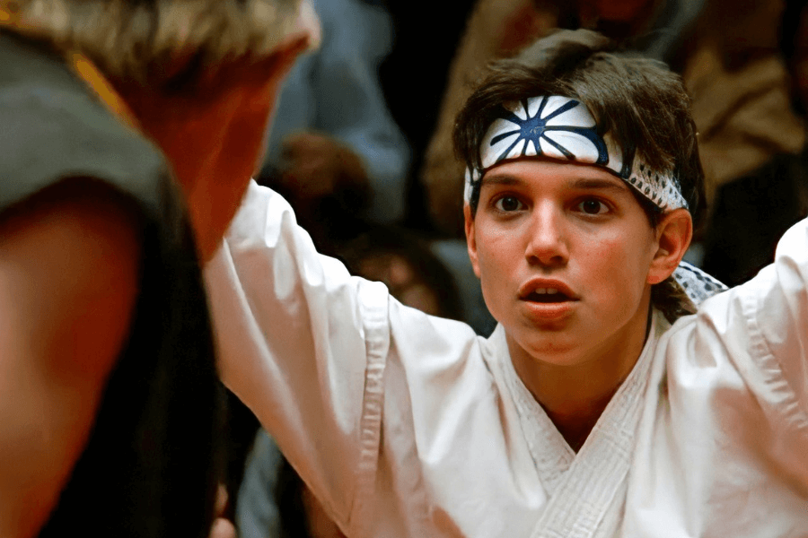 This image shows a young man in a white martial arts uniform and a headband with a sunburst design, looking focused and determined. He appears to be engaged in a karate match or training, with his arms raised. The background is blurred, depicting spectators or a crowded event, adding an intense and competitive atmosphere to the scene.