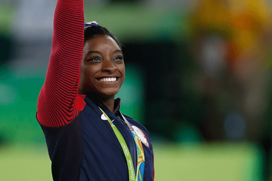 A female athlete with a bright smile raises her arm in victory while wearing a Team USA tracksuit. A gold medal hangs around her neck, and the background shows a blurred Olympic setting with green and yellow hues. Her expression radiates pride and joy.